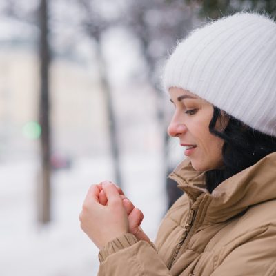 A young girl in a white hat warms her hands. It stands in the city in winter under the snow